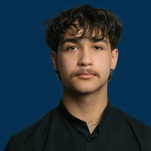 Alexander vom Aveda Exclusive Salon in Zürich Enge, mit dunklem lockigem Haar und Schnurrbart, im schwarzen Hemd vor dunkelblauem Hintergrund