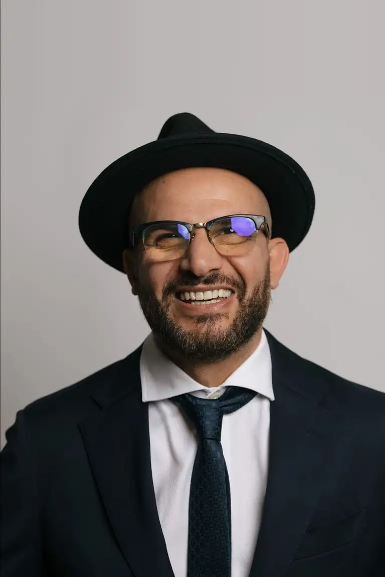 Eddine Belaid lachend, mit Hut und Brille im dunklen Anzug, Porträt im Hochformat vor hellgrauem Hintergrund